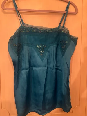 Turkos satin linnetopp med paljetter - Snygg turkos linnetopp i satin med smala axelband och spetsdetaljer upptill. Toppen har paljetter längs kanten och ett glansigt material som ger en lyxig känsla. Perfekt att styla med jeans eller kjol för en trendig look.