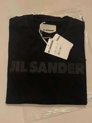 Svart Jil Sander t-shirt med logga - Stilren svart t-shirt från Jil Sander med stor logga tryckt framtill. Klassisk passform och rund halsringning. Tillverkad i mjuk bomull för en skön känsla. Perfekt för dig som gillar minimalistisk design och premiumkänsla.