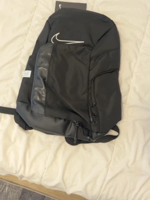 Svart Nike Elite ryggsäck - Snygg svart Nike Elite ryggsäck med vit logga och detaljer. Ryggsäcken har flera fack, vadderade axelband med silvriga detaljer och justerbar bröstrem. Tillverkad i slitstarkt syntetmaterial och passar perfekt för skola, träning eller cityhäng.