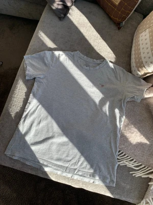 Grå t-shirt från Gant - En enkel grå t-shirt från Gant med liten röd logga på bröstet. Klassisk rund hals och korta ärmar. Perfekt till jeans eller shorts och gjord i mjuk bomull för skön känsla hela dagen.