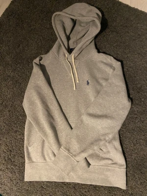 Grå hoodie från Polo Ralph Lauren - Snygg grå hoodie från Polo Ralph Lauren med klassisk huva och dragsnören. Broderad blå logga på bröstet och stor känguruficka framtill. Mjuk bomullsblandning som är skön att ha på sig. Perfekt för en avslappnad stil. Det är tre fläckar på ryggen men inget som märks när man har på den