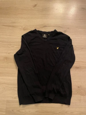 Svart stickad tröja från Lyle & Scott - Svart stickad tröja från Lyle & Scott med V-ringning och gul broderad logga på bröstet. Tröjan har långa ärmar och ribbade muddar vid ärmslut och nederkant. Perfekt för lager-på-lager och enkel att matcha. Säljer pga ingen användning och för att den inte passar mig längre!