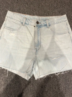 Ljusblå denimshorts - Säljer ett par ljusblå denimshorts från 4Denim med råa, fransiga benslut och slitna detaljer framtill. Klassisk femficksmodell med knapp och dragkedja. Perfekta för varma dagar och enkel att matcha med allt.