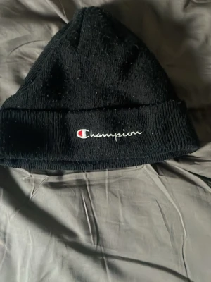 Svart ribbad mössa från Champion - Säljer en svart ribbstickad mössa från Champion med uppvikt kant och broderad logga framtill. Klassisk och enkel design som passar perfekt till kalla dagar. Materialet känns mjukt och bekvämt mot huvudet.