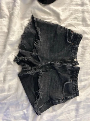 Svarta jeansshorts med fransig kant - Snygga svarta jeansshorts med hög midja och rå, fransig kant. Klassisk femficksmodell med dragkedja och knapp framtill. Perfekta för en avslappnad och trendig look.