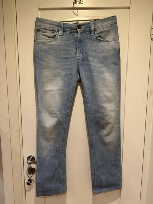 Nudie jeans  - Säljer ett par ljusblå jeans från Nudie med raka ben och klassisk femficksmodell. Jeansen har diskreta slitningar och en snygg tvättad look. Tillverkade i bomull med en bekväm passform, perfekta för en avslappnad stil.