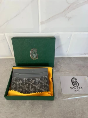 Korthållare - Snygg grå korthållare från Goya** med klassiskt mönster i vitt och svart. Tillverkad i canvas och läder, med flera fack för kort. Kommer i original grön ask och med gul putsduk samt Goy*** Paris-kort. Perfekt accessoar för dig som gillar exklusiva detaljer .  Pris kan ev disskuteras, enbart seriösa köpare. Kan vid efterfrågan även mötas i Stockholm. Allt är i nyskick