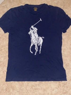 Marinblå t-shirt från Polo Ralph Lauren - Snygg marinblå t-shirt från Polo Ralph Lauren med det klassiska stora polospel-trycket i vitt på bröstet. T-shirten har rund hals och korta ärmar, perfekt för en avslappnad och stilren look.