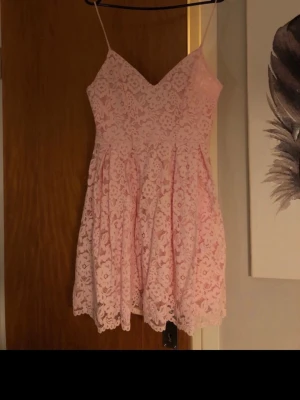 Rosa spetsklänning med smala axelband - Söt rosa kortklänning med smala axelband och v-ringad urringning. Klänningen har ett vackert spetsmönster över hela plagget och en lätt utställd kjol. Perfekt för sommarens alla tillfällen.