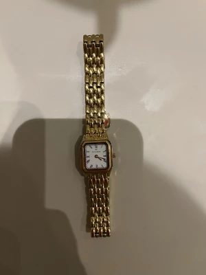 Guldfärgad klocka Mockberg - Stilren guldfärgad klocka från Coach med fyrkantig vit urtavla och länkarmband i metall. Klassisk design med tydliga svarta siffror och röda detaljer på visarna. Perfekt accessoar för dig som gillar tidlös stil.