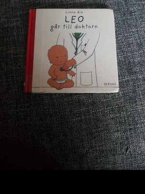 Leo går till doktorn av Linne Bie - En barnbok med hårda pärmar, 'Leo går till doktorn' av Linne Bie. Omslaget är vitt med röd rygg och visar en illustration av en bebis och en läkare. Perfekt för små barn som vill läsa om vardagliga situationer.