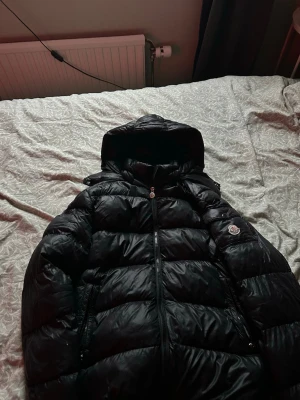 Svart dunjacka med huva från Moncler - Säljer en svart dunjacka från Moncler med huva och dragkedja framtill. Jackan har en ficka med knapp och Moncler-logga på ärmen. Den är quiltad och har en glansig finish som ger en riktigt snygg look. Perfekt för kalla vinterdagar. Den har en liten defekt som inte syns alls nästan. Byten är intressant så bara visa mig vad du är villig att byta söker helst flycrews eller lanvins 