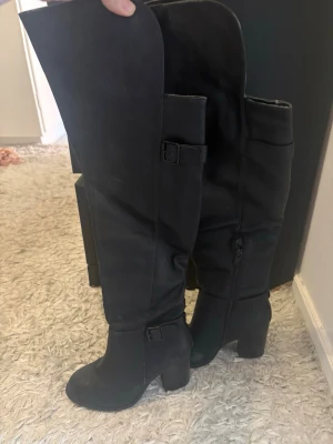 Svarta overknee stövlar med klack - Säljer ett par svarta overknee stövlar med chunky klack och dekorativa spännen. Stövlarna har dragkedja på insidan och är tillverkade i ett matt material som liknar mocka. Perfekta för att lyfta vilken outfit som helst.