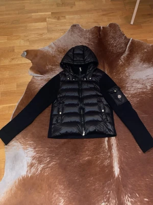 Svart dunväst med huva från Moncler - Snygg svart dunväst från Moncler med huva och stickade ärmar. Västen har två fickor med tryckknappar och en ikonisk Moncler-logga på ärmen. Materialet är glansigt dun på kroppen och ribbstickat tyg på ärmarna. Perfekt för lager-på-lager och streetstyle.