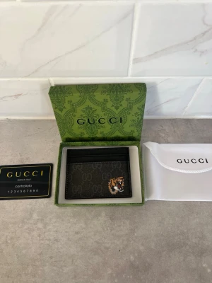 Korthållare - Snygg svart korthållare från GG i canvas och läder med klassiskt GG-mönster och en cool tigerpatch på framsidan. Kommer i original grön ask och med dustbag samt äkthetskort. Perfekt för dig som vill ha något stilrent men ändå unikt. Möts ev i stockholm också.