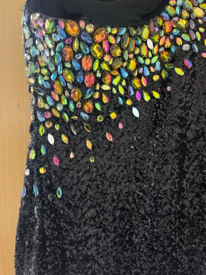 Svart glittrig oneshoulderklänning - Svart oneshoulderklänning täckt med svarta paljetter och stora färgglada stenar upptill. Klänningen har en asymmetrisk axel och är figurnära. Perfekt för dig som vill sticka ut med en unik och glittrig look.