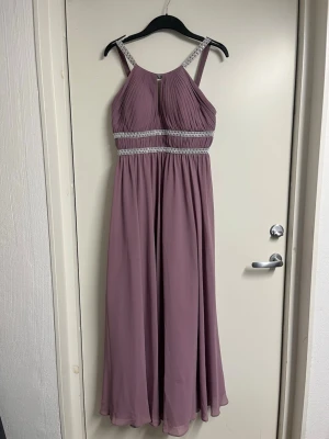 Lång lila klänning med smala band - Elegant långklänning i lila med smala axelband och dekorativa band med silverdetaljer vid midjan och axelpartiet. Klänningen har ett plisserat liv och faller mjukt från midjan ner. Perfekt för speciella tillfällen.