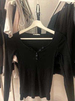 Svart långärmad topp med knappar - Snygg svart långärmad topp med v-ringning och dekorativa knappar framtill. Modellen är figurnära och passar perfekt till jeans eller kjol. Tillverkad i mjukt material som känns skönt mot huden.