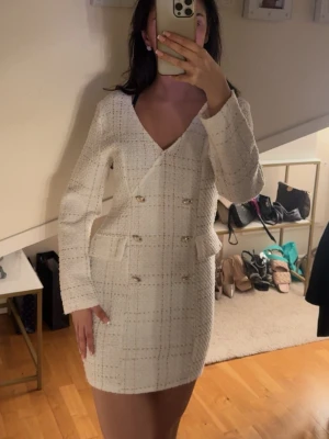 Elegant klänning  - Stilren vit klänning i rutigt mönster med dekorativa silvriga knappar framtill. Klänningen har v-ringning, lång ärm och markerad midja. Materialet är strukturerat och ger en exklusiv känsla. Perfekt för dig som gillar klassisk och trendig stil.