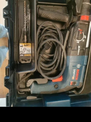 Blå Bosch slagborrmaskin i väska - Säljer en blå och svart Bosch slagborrmaskin med lång sladd, extra handtag och flera borr i originalväska. Perfekt för dig som gillar att fixa hemma eller behöver ett kraftfullt verktyg till byggprojekt. Allt ligger smidigt packat i en robust plastväska.