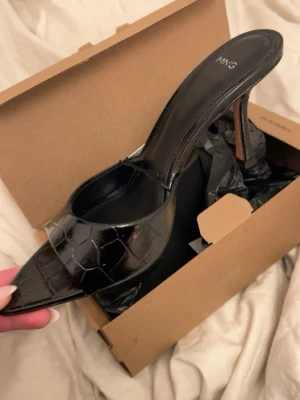 Svarta pumps med krokopräglad yta från MNG - Säljer ett par svarta pumps från MNG med spetsig tå och hög klack. Skorna har en snygg krokopräglad yta i konstläder och öppen baksida. Perfekta för dig som vill ha en trendig och elegant look.