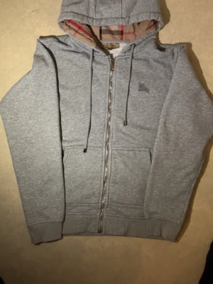 Grå zip hoodie från Burberry - Snygg grå hoodie från Burberry med dragkedja, fickor fram och klassiskt rutigt foder i huvan. Tillverkad i mjukt material och har justerbara snören i huvan. Perfekt för en avslappnad och stilren look. PRISET KAN DISKUTERAS