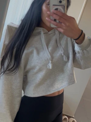 Grå croppad hoodie med huva - Säljer en ljusgrå croppad hoodie med huva och dragsnören. Hoodien har långa ärmar och en avslappnad passform, perfekt för en chill och trendig look. Materialet är mjukt och skönt, troligtvis bomullsblandning. Inga synliga loggor eller märken.