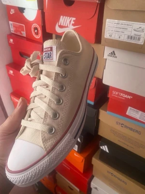 Beige Converse All Star låga sneakers - Säljer ett par klassiska Converse All Star sneakers i beige canvas med vita gummisulor och röda detaljer. Skorna har lågt skaft, vita snören och metallöljetter. Perfekta för en avslappnad och stilren look.