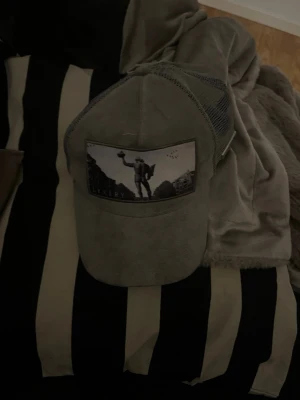 Grå keps från LYXERY BY SWEDEN - Snygg grå keps från LYXERY BY SWEDEN med mesh på sidorna och justerbar rem bak. Framtill finns ett tryck med motiv av en person och landskap. Insidan har svartvitt kartmönster och band med logotyp. Perfekt för en cool och avslappnad stil.