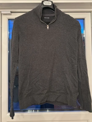 Mörkgrå halvzip tröja Jack & Jones - Snygg mörkgrå tröja från Jack & Jones med halvzip och hög krage. Tillverkad i mjukt material, perfekt för lager på lager. Tröjan har långa ärmar och ribbade muddar vid ärmslut och nederkant. Enkel och stilren design som passar till det mesta.