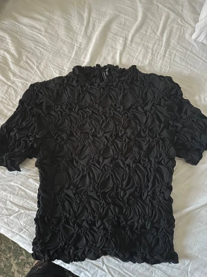 Svart topp  - Cool svart topp från Vero Moda med unik twistad och rynkad struktur över hela plagget. Kortärmad modell med högre halsringning och stretchigt material som ger en snygg passform. Perfekt för dig som vill sticka ut lite extra.