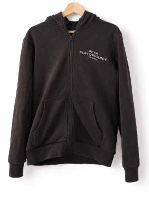 Svart hoodie Peak Performance - Svart hoodie med dragkedja från Peak Performance. Klassisk modell med huva, två stora fickor fram och ribbade muddar. Diskret logga i vitt på bröstet. Tillverkad i mjukt bomullsmaterial som känns skönt mot huden.