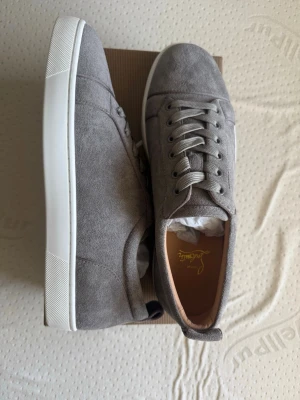 Gråa mockasneakers från Christian Louboutin - Snygga sneakers i grå mocka från Christian Louboutin med vita sulor och matchande grå skosnören. Klassisk låg modell med diskret design och exklusiv känsla. Perfekta för dig som gillar stilrena och lyxiga detaljer.