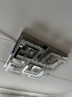 Modern taklampa i kristall - Säljer en fyrkantig taklampa i modern design med flera lager av kromade ramar och gnistrande kristalldetaljer. Lampan har geometriska former och ger ett lyxigt intryck till rummet. Perfekt för dig som gillar stilren och exklusiv inredning. Lampan är kopplad till en fjärrkontroll som ändrar färgen men även hur stark lampan ska lysa! 