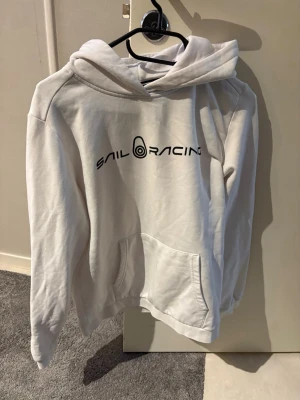Vit hoodie från Sail Racing - Säljer en vit hoodie från Sail Racing med svart logga tryckt på bröstet. Hoodien har en klassisk huva och en stor magficka framtill. Tillverkad i mjukt material som känns skönt mot huden. Perfekt för dig som gillar enkel och stilren design.