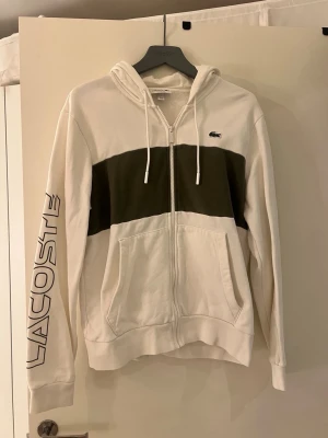 Vit hoodie med dragkedja från Lacoste - Snygg vit hoodie från Lacoste med dragkedja, huva och snören. Stor grön panel över bröstet och stor Lacoste-logga längs ena ärmen. Klassisk krokodil-logga på bröstet. Två fickor framtill och ribbade muddar. Perfekt för en sportig och avslappnad stil.