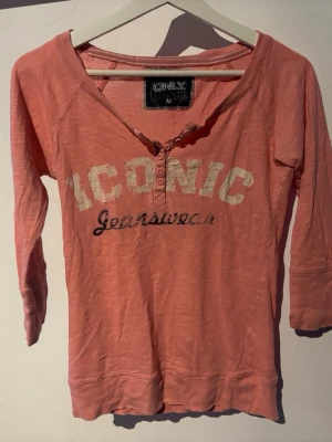 Korallrosa topp från ONLY med tryck - Säljer för den e för liten( hel ny) Korallrosa topp från ONLY med texten 'ICONIC Jeanswear' framtill. Toppen har v-ringning med knappar och trekvartsärm. Tillverkad i mjuk bomull, perfekt för en avslappnad look.