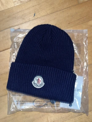 Mörkblå Moncler mössa i ull - Snygg mörkblå ribbstickad mössa från Moncler med uppvikt kant och klassisk Moncler-logga framtill. Tillverkad i mjuk ull som håller dig varm och ger en clean look. Perfekt accessoar för kalla dagar.