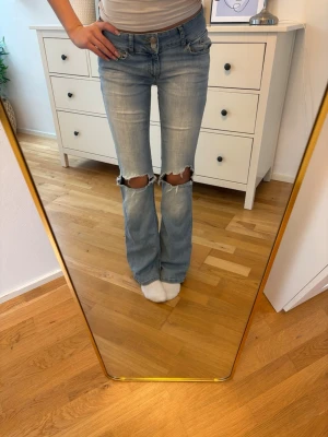 Ljusblå slitna bootcut jeans från Nelly  - Säljer ett par ljusblå bootcut jeans från Nelly med stora hål över knäna och klassisk femficksmodell. Jeansen har låg midja och är tillverkade i mjuk denim med lite stretch för skön passform. Perfekta för en avslappnad och trendig look. Säljer då jeansen är för stora!! 