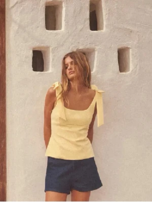 Gul linnetopp med knytband - Söt gul linnetopp med breda axelband som knyts i rosetter på axlarna. Toppen har en rak, något figurnära passform och är perfekt för varma dagar. Tillverkad i ett lätt material som känns luftigt och somrigt.