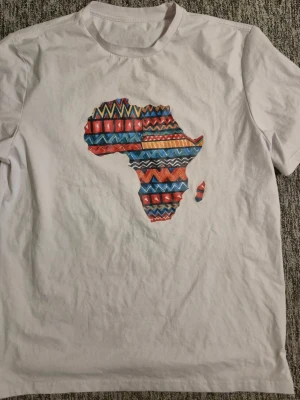 Vit t-shirt med färgglad Afrika-tryck - Vit t-shirt med ett stort tryck av Afrikas kontinent på bröstet, dekorerad med färgglada geometriska mönster i rött, blått, gult och svart. Klassisk rund hals och korta ärmar. Perfekt för dig som vill sticka ut med en unik och kreativ design. Storlek XL/XXL, finns i hässelby eller skickas. 