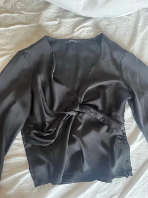 Svart topp från Gina - Snygg svart omlottopp från Ginatricot i glansigt satinmaterial. Toppen har långa ärmar, v-ringning och en knytning framtill som ger en cool twist. Nästan aldrig använd eftersom jag tycker den är lite stor på mig! 💕