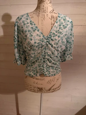 Grönblommig v-ringad blus - Söt blus med grönt bladmönster på vit botten. V-ringad framtill med dekorativa knappar och rynkdetaljer längs mitten. Korta puffärmar med volangkant och lätt figurnära passform. Tunn och luftig kvalitet, perfekt för vår och sommar.