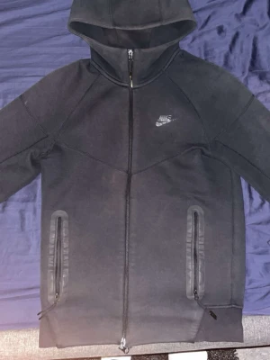 Svart Nike tech fleece med dragkedja - Snygg svart Nike tech fleece från Nike med hel dragkedja framtill och två fickor med dragkedja. Klassisk Nike-logga på bröstet och huva för extra stilpoäng. Tillverkad i mjukt och bekvämt material, perfekt för en avslappnad streetwear-look.