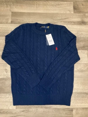 Mörkblå kabelstickad tröja Polo Ralph Lauren - Mörkblå kabelstickad tröja från Polo Ralph Lauren med rund halsringning och röd broderad logga på bröstet. Tröjan har ribbade muddar vid ärmslut och nederkant. Klassisk och tidlös design i mjukt material.