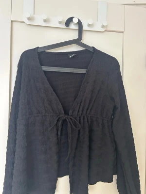 Svart omlottopp från Gina Tricot - Säljer en svart omlottopp från Gina Tricot i stretchigt material. Toppen har lång ärm, v-ringning och knyt framtill i midjan. Perfekt att styla med jeans eller kjol för en trendig look.