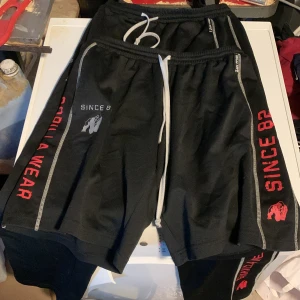 Svarta Gorilla Wear träningsbyxor och shorts  - Säljer ett par svarta byxor och shorts  från Gorilla Wear med vita dragsnören och tryck i rött och vitt längs benen. Texten 'SINCE 82' och 'SPORTSWEAR' syns tydligt, samt Gorilla Wear-loggan. Perfekta för gymmet eller chill hemma. På shortsen står storleken L/XL och byxorna S/M men skulle säga att båda är M/L. Passar mig bra som är 175 75kg. Gymbyxor