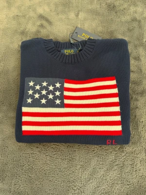 Mörkblå stickad tröja Polo Ralph Lauren - Mörkblå stickad tröja från Polo Ralph Lauren med amerikansk flagga i rött, vitt och blått på bröstet. Rund halsringning och ribbstickade muddar. Klassisk och stilren design med RL-broderi i hörnet. Perfekt för dig som gillar preppy stil.