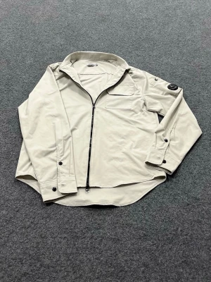 C.P Company Cardigan - Stilren beige cardiganjacka från C.P. Company med dragkedja framtill, bröstficka och klassisk linsdetalj på ärmen. Jackan har en avslappnad passform och är tillverkad i ett mjukt, lätt material som passar perfekt till lager-på-lager.