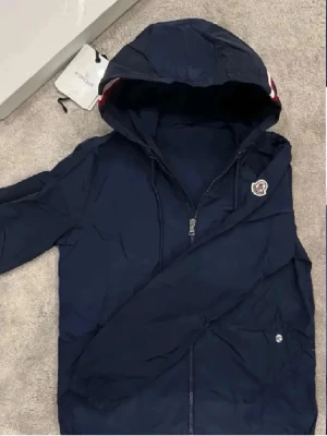 Mörkblå vindjacka från Moncler - Snygg mörkblå vindjacka från Moncler med huva och dragkedja. Jackan har Moncler-logga på bröstet och en röd, vit och blå rand på huvan. Tillverkad i lätt, glansigt material som står emot vind. Perfekt för en sportig och stilren look. Lappar ingår ej